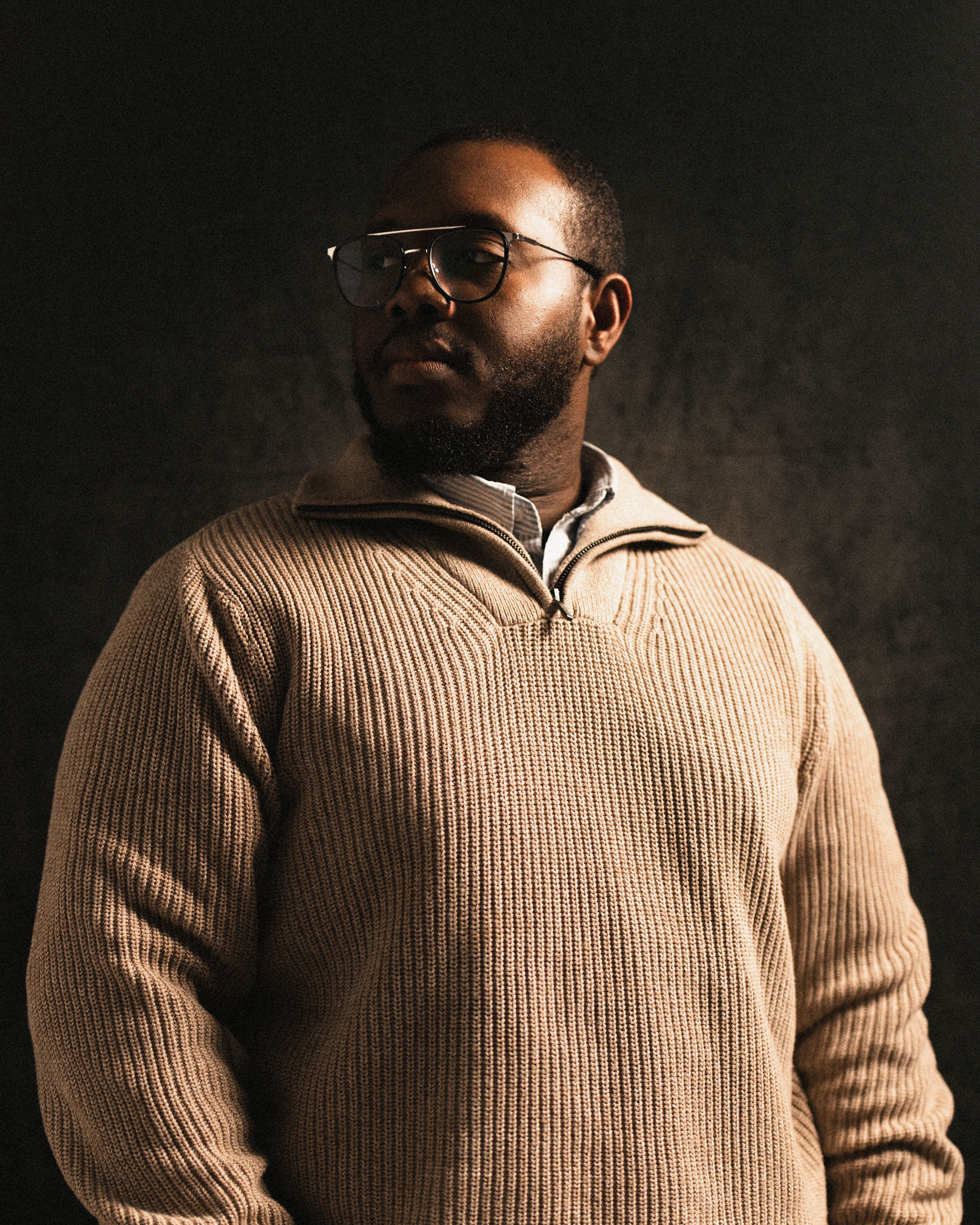 A reflective portrait of a man wearing glasses and a ribbed sweater against a dark backdrop.