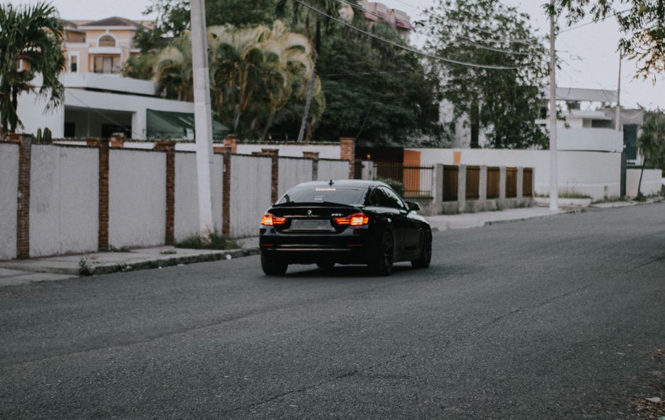 Modern black passenger car with glowing taillights driving through deserted suburb street with high green trees on roadside