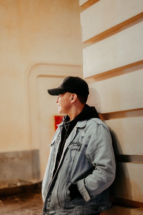 Free Casual young man in denim jacket and cap leaning against a wall. Urban indoor setting. Stock Photo