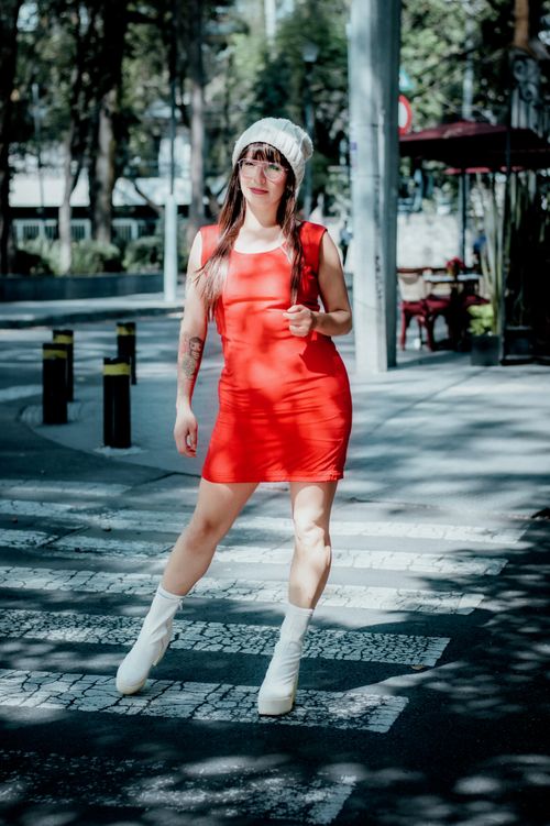 Free Young woman in red dress and white boots crossing a city street with sunlight filtering through trees. Stock Photo
