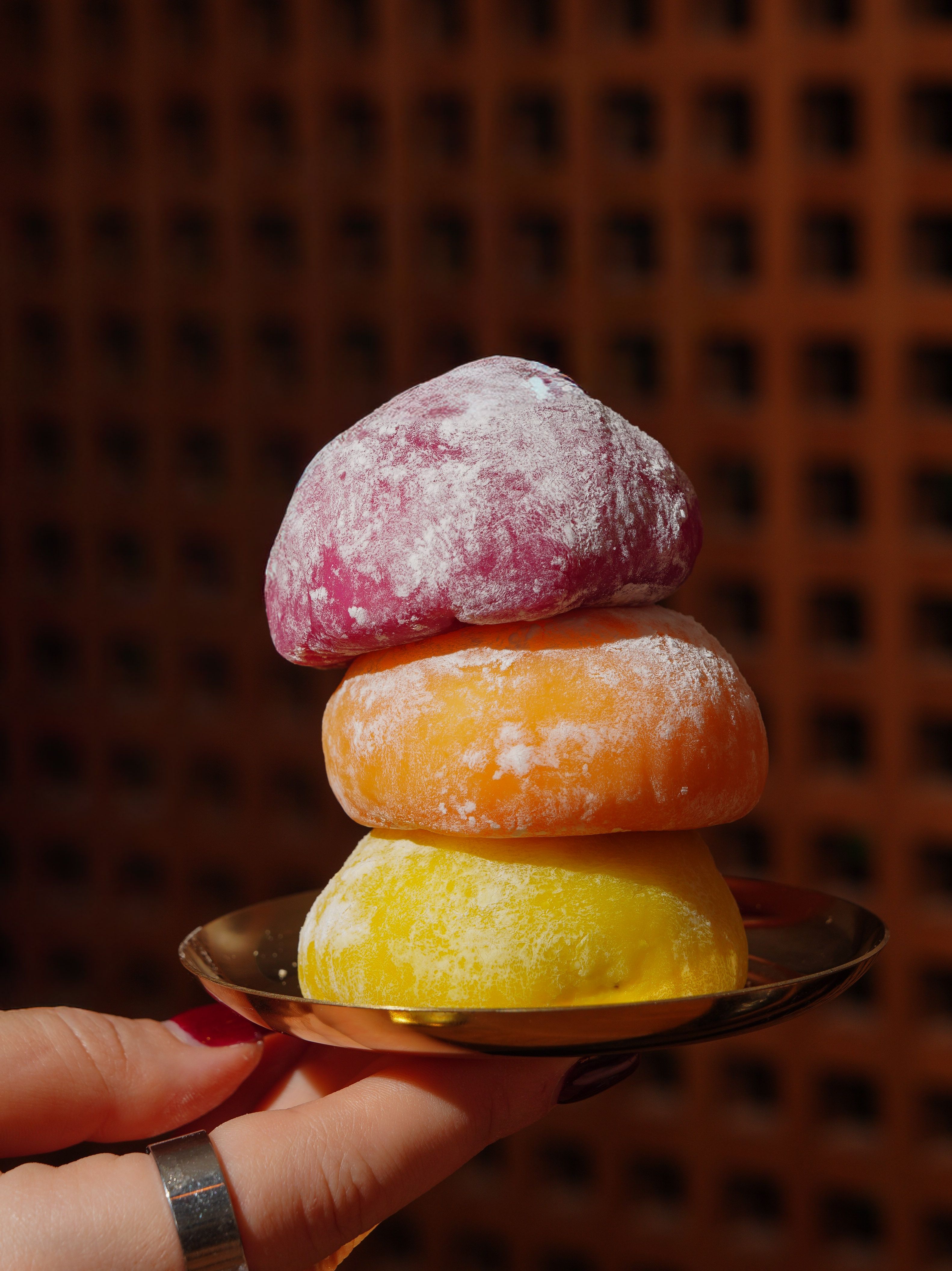 Three colorful mochi desserts stacked on a gold plate outdoors, showcasing vibrant colors and textures.