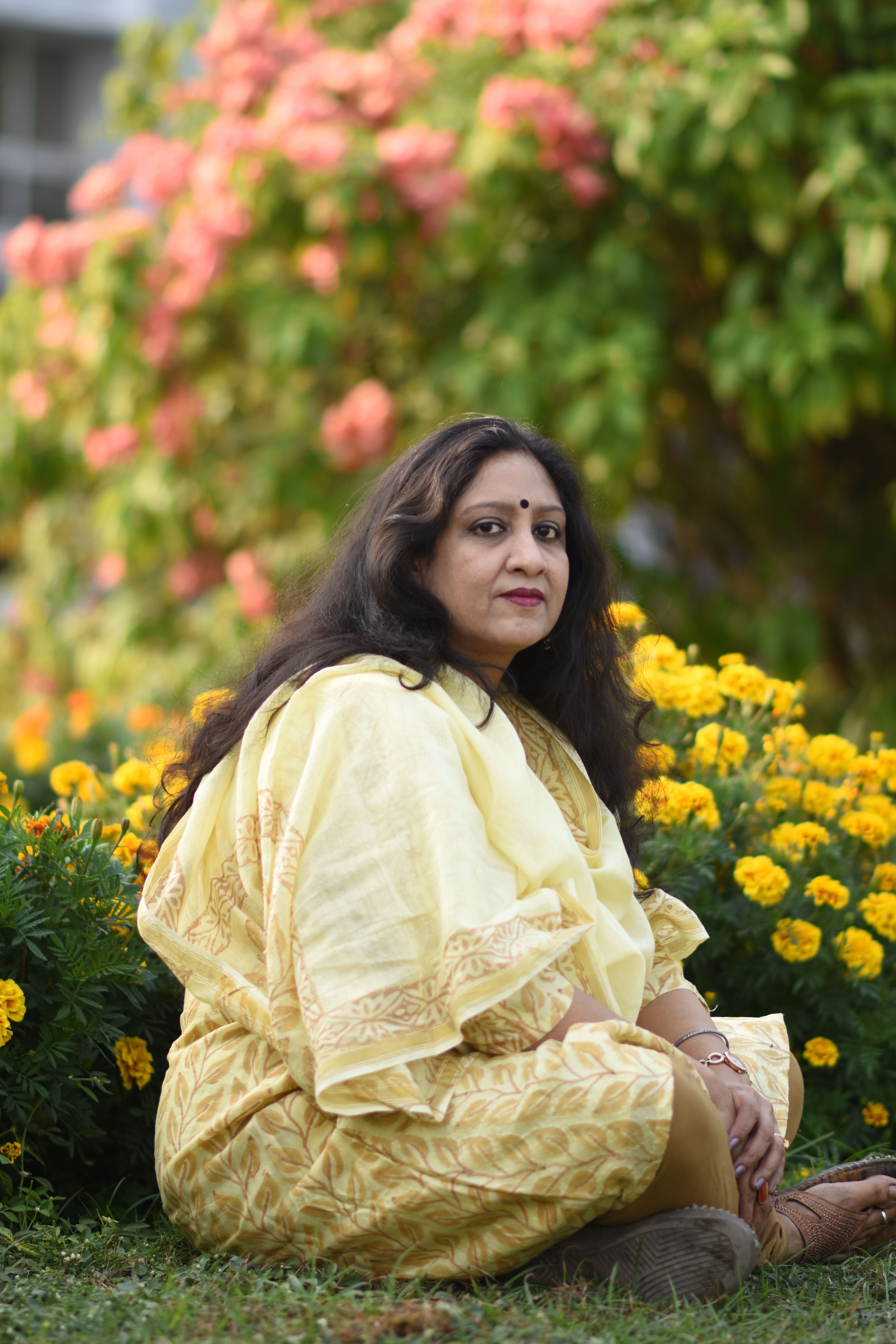 A woman in a yellow sari sits gracefully in a colorful garden, surrounded by vibrant blooms.