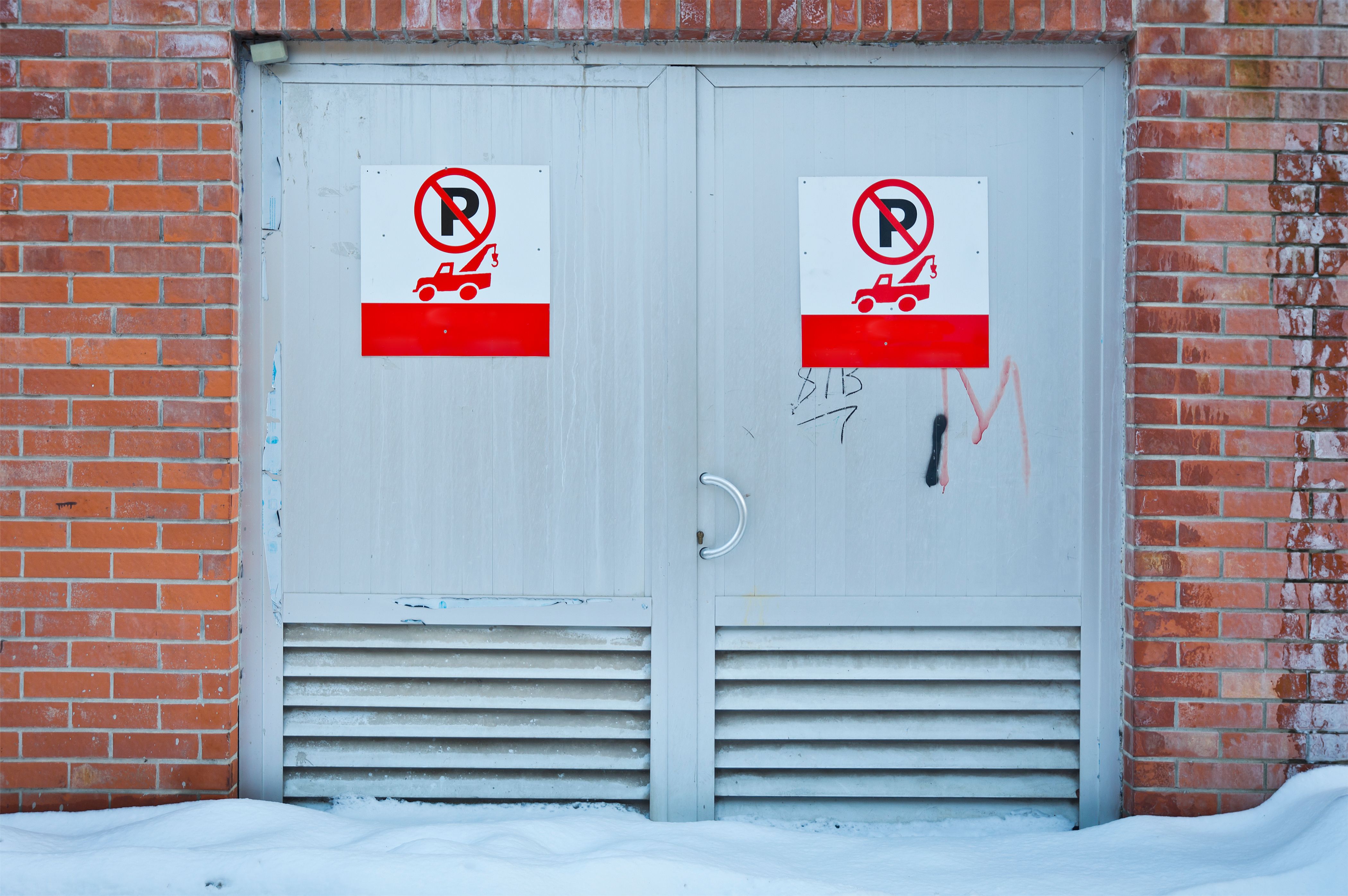 Front view of no parking signs on a metal door framed by a brick wall, with snow on the ground.