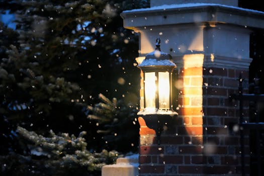 A glowing lantern on a brick wall reflects warm light amidst falling snowflakes on a quiet winter evening.