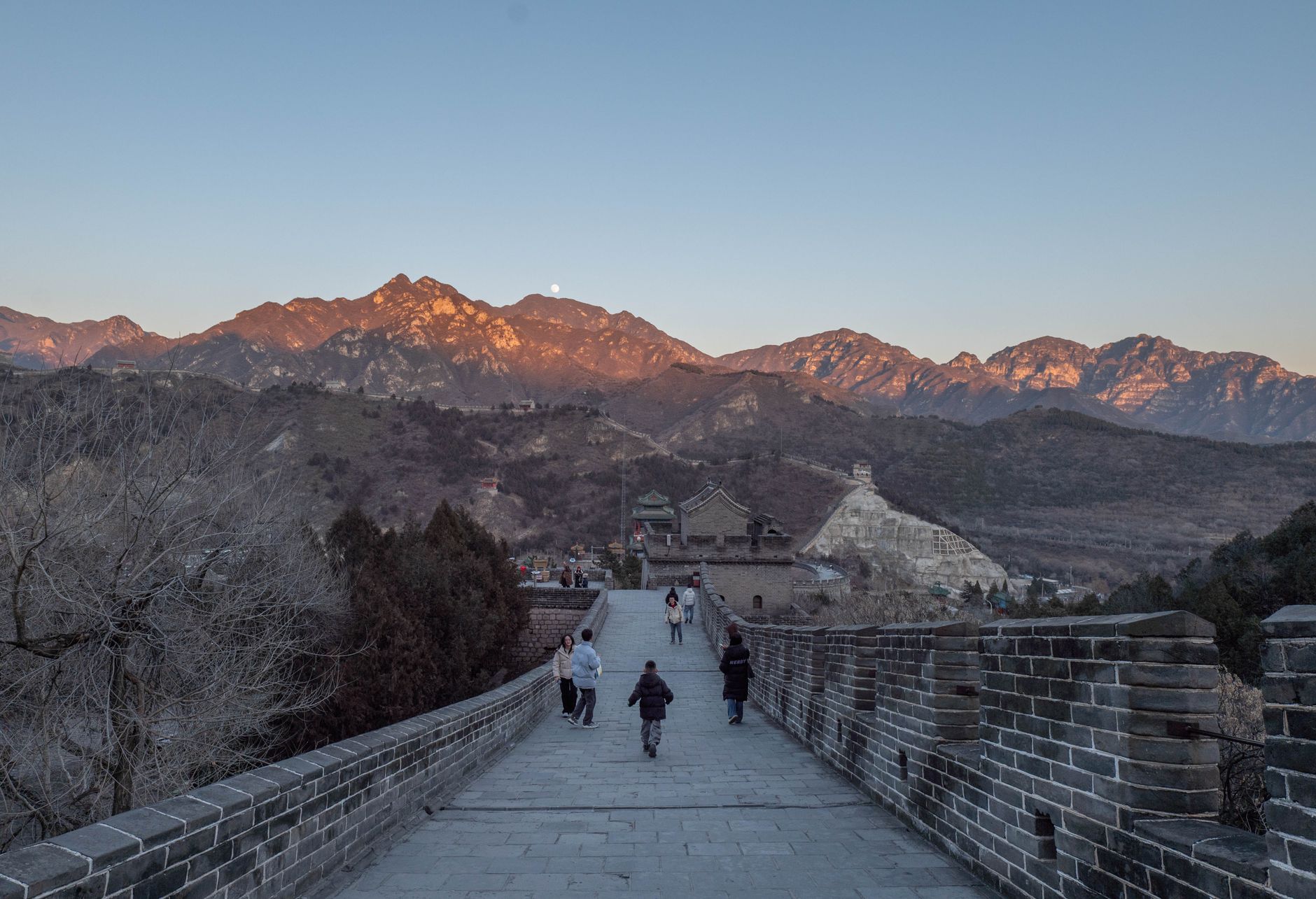 Tourists walking on the Great Wall of China at sunset with mountains in the background.