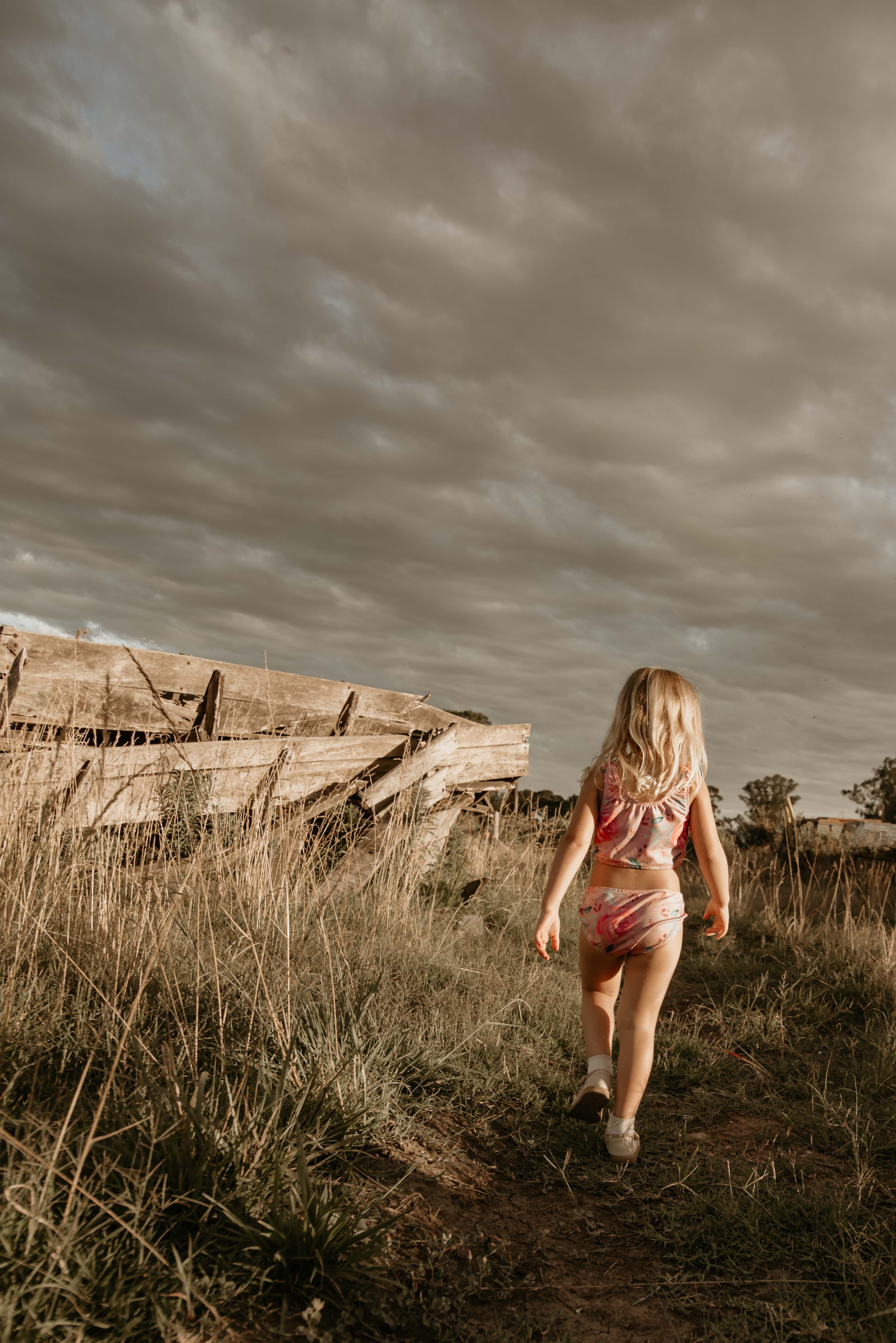 A child walks near a rustic wooden fence in an open field under a dramatic cloudy sky.