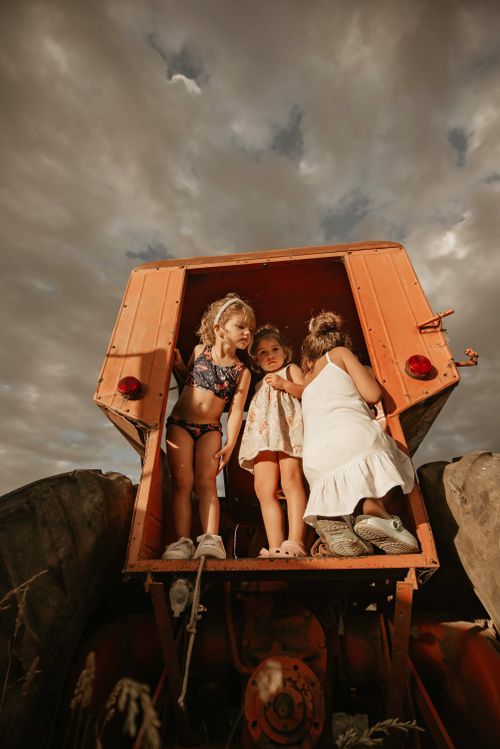 Free Three children playing on a rustic red tractor in an open field under cloudy skies. Stock Photo