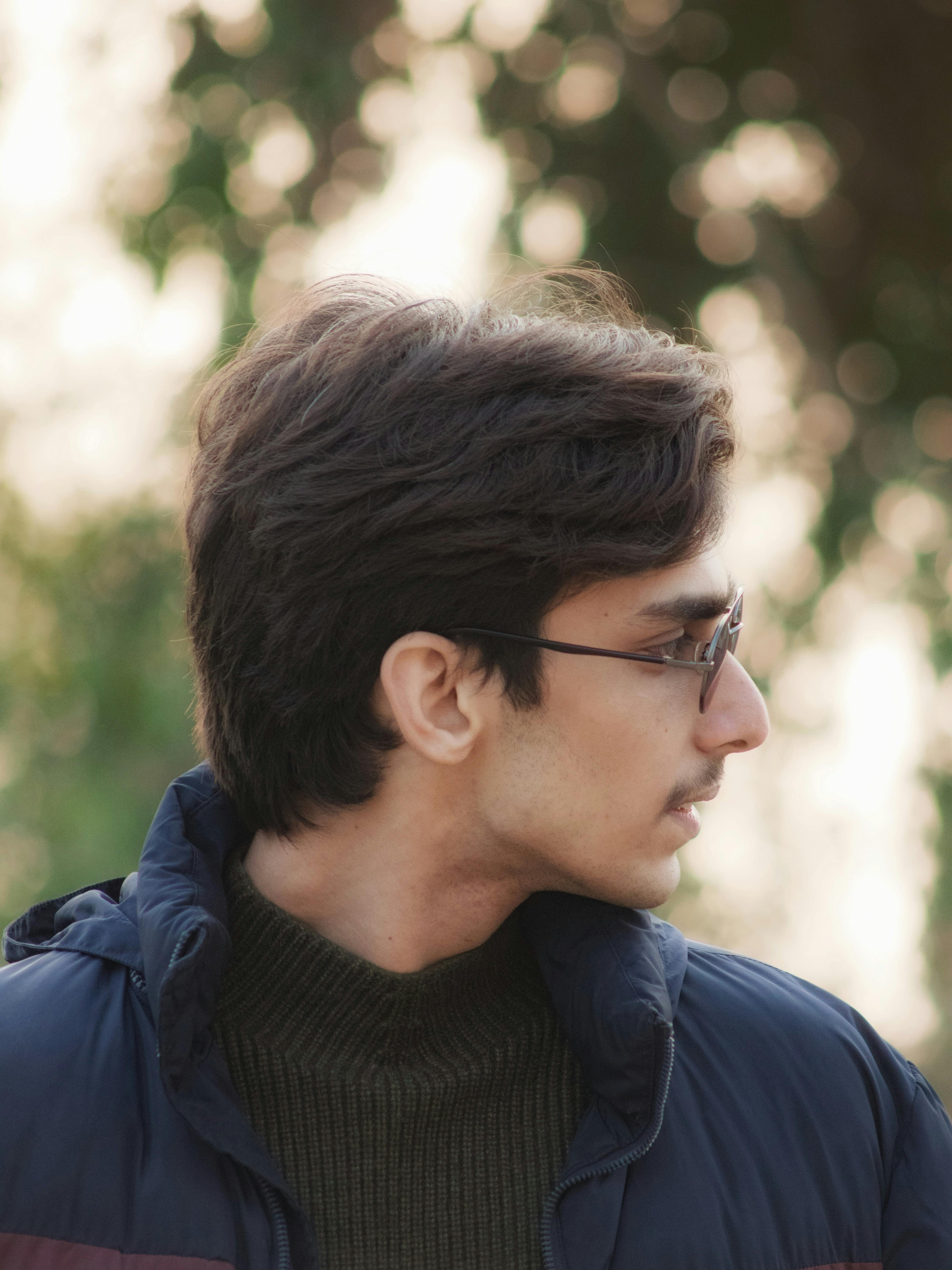 A thoughtful portrait of a young man wearing glasses and a winter coat, set against a blurred outdoor backdrop.