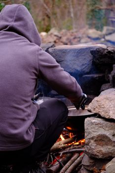 A person in a hoodie cooks over a campfire surrounded by stones, creating a warm atmosphere.