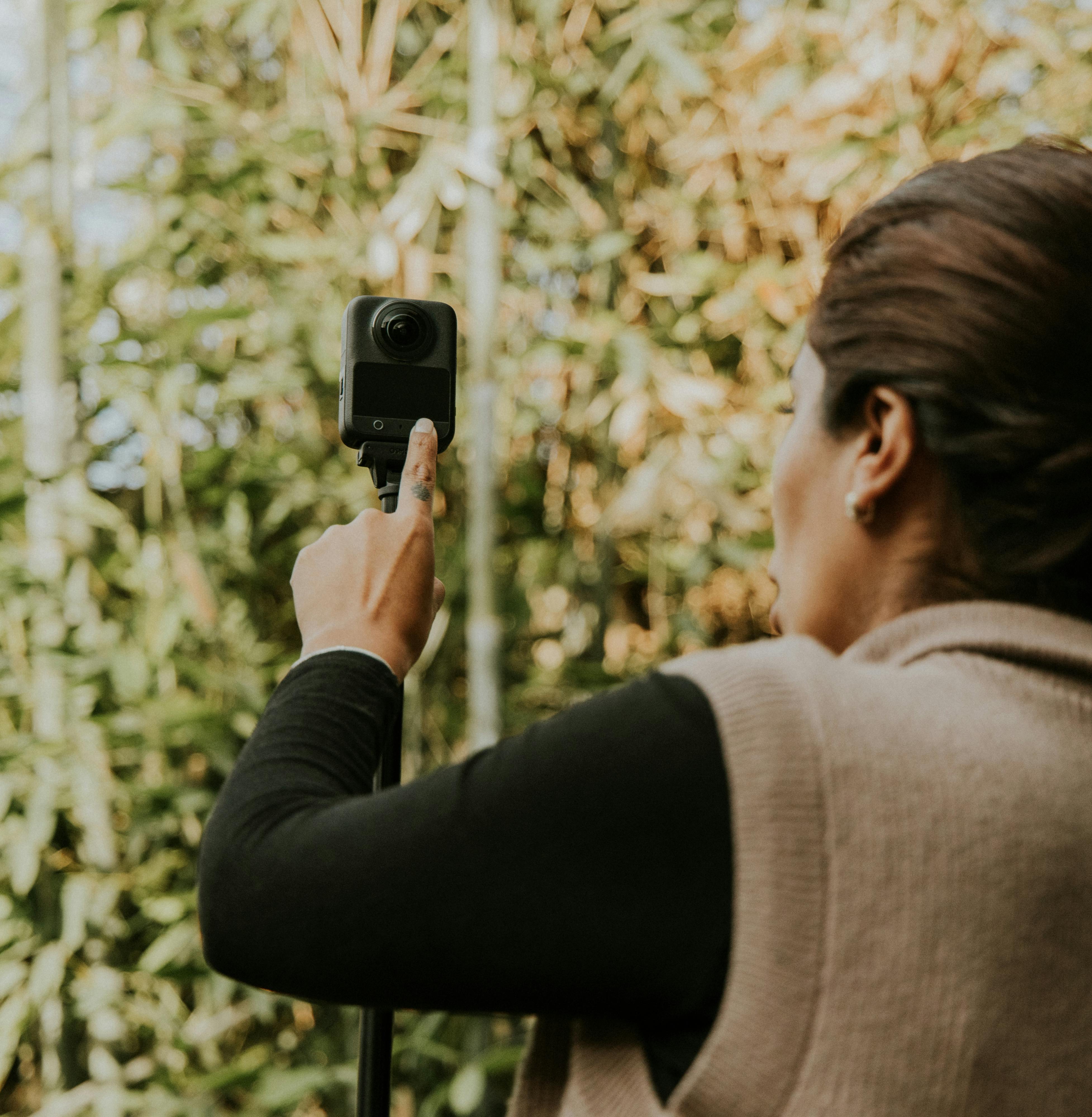 Free Rear view of a woman capturing video with a 360 camera outdoors amidst greenery. Stock Photo