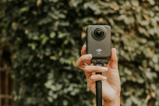 Detailed shot of a hand holding a 360 camera against a leafy blurred background.