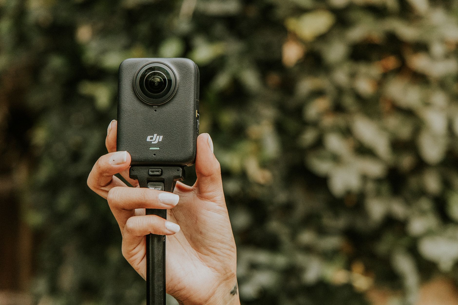 Close-up of a hand holding a modern 360-degree camera against a blurred green background.