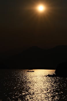 A boat in silhouette casts a reflection on shimmering water under a golden sunset.