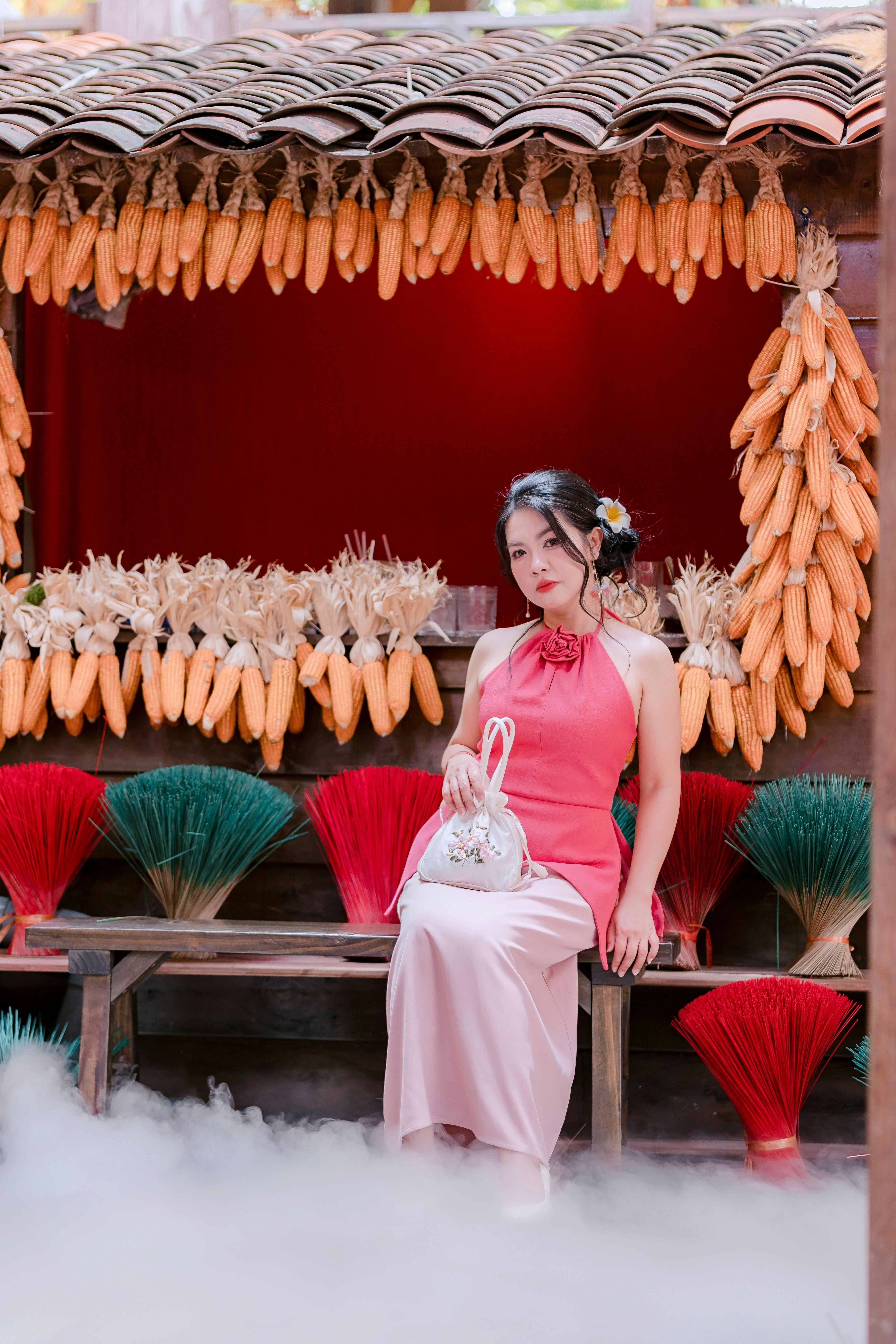 Woman in a red dress sitting by colorful brooms at a corn-adorned stall.