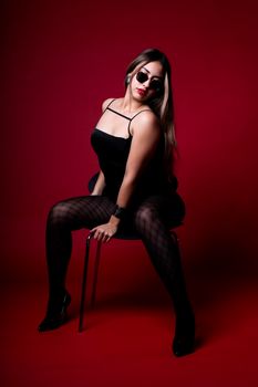 Stylish woman in black attire sitting confidently against a vibrant red backdrop.