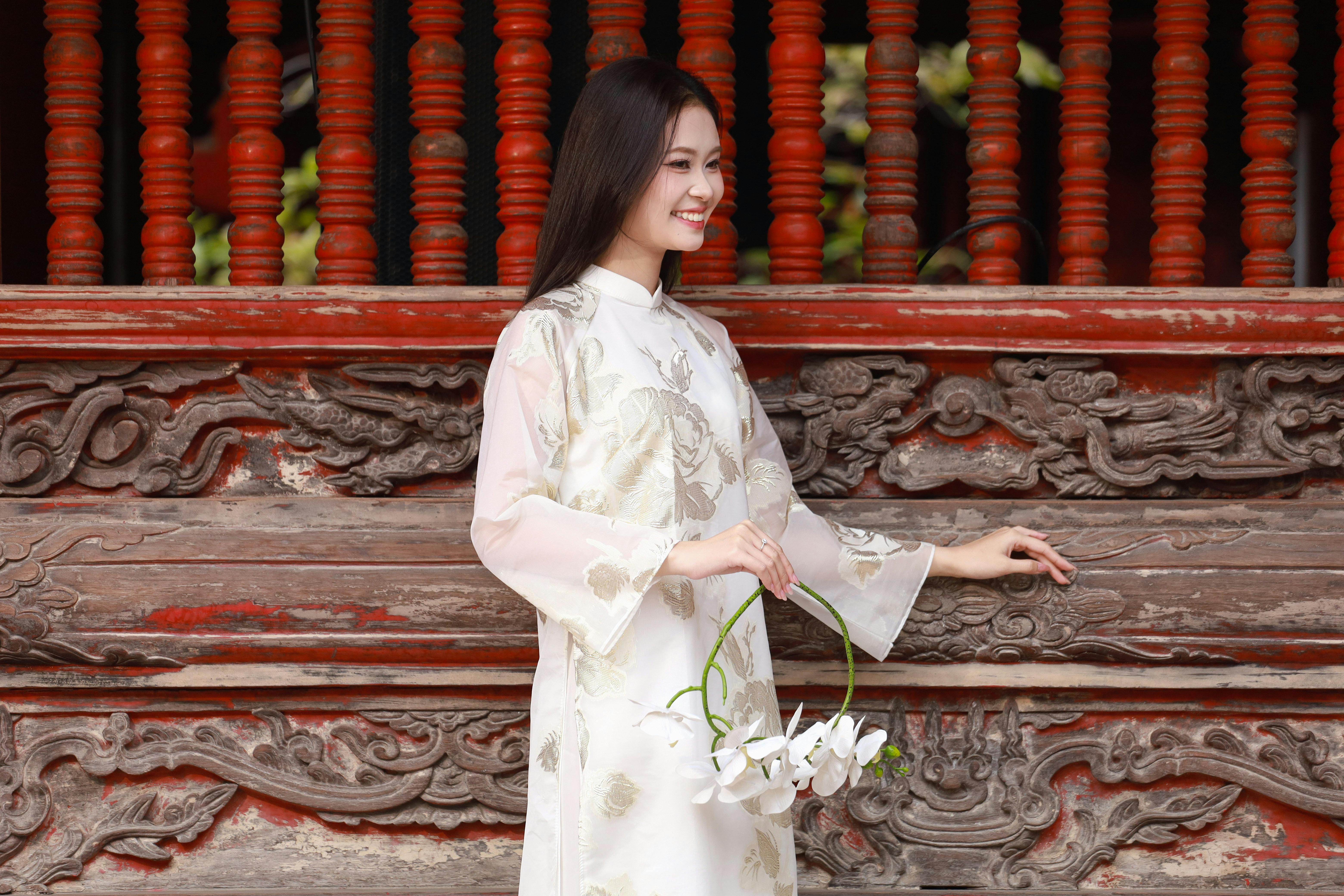 grátis Jovem mulher vestida com um tradicional áo dài vietnamita, segurando flores de lótus e sorrindo contra um painel de madeira ornamentado. Foto profissional