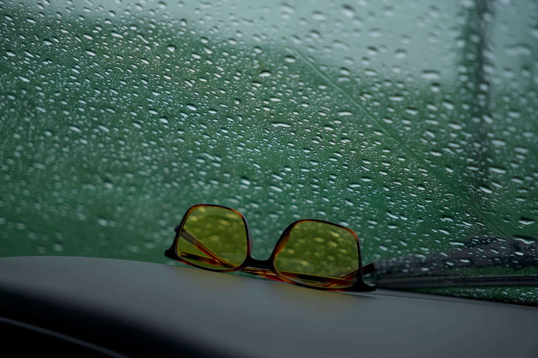 Yellow sunglasses rest on a car dashboard against a rain-speckled window.