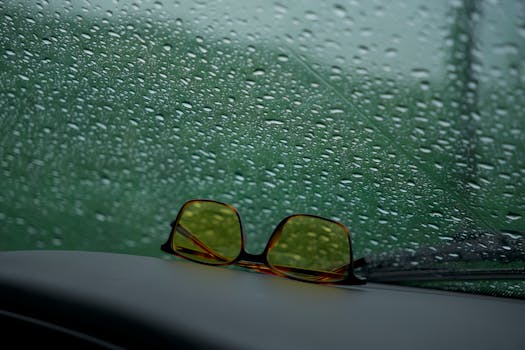 Yellow sunglasses rest on a car dashboard against a rain-speckled window.