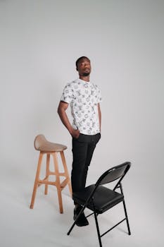 Man stands in studio wearing printed shirt, with a stool and chair beside him.