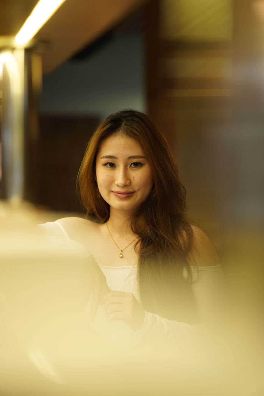 Portrait of a young woman with long hair, smiling in a softly lit indoor setting.