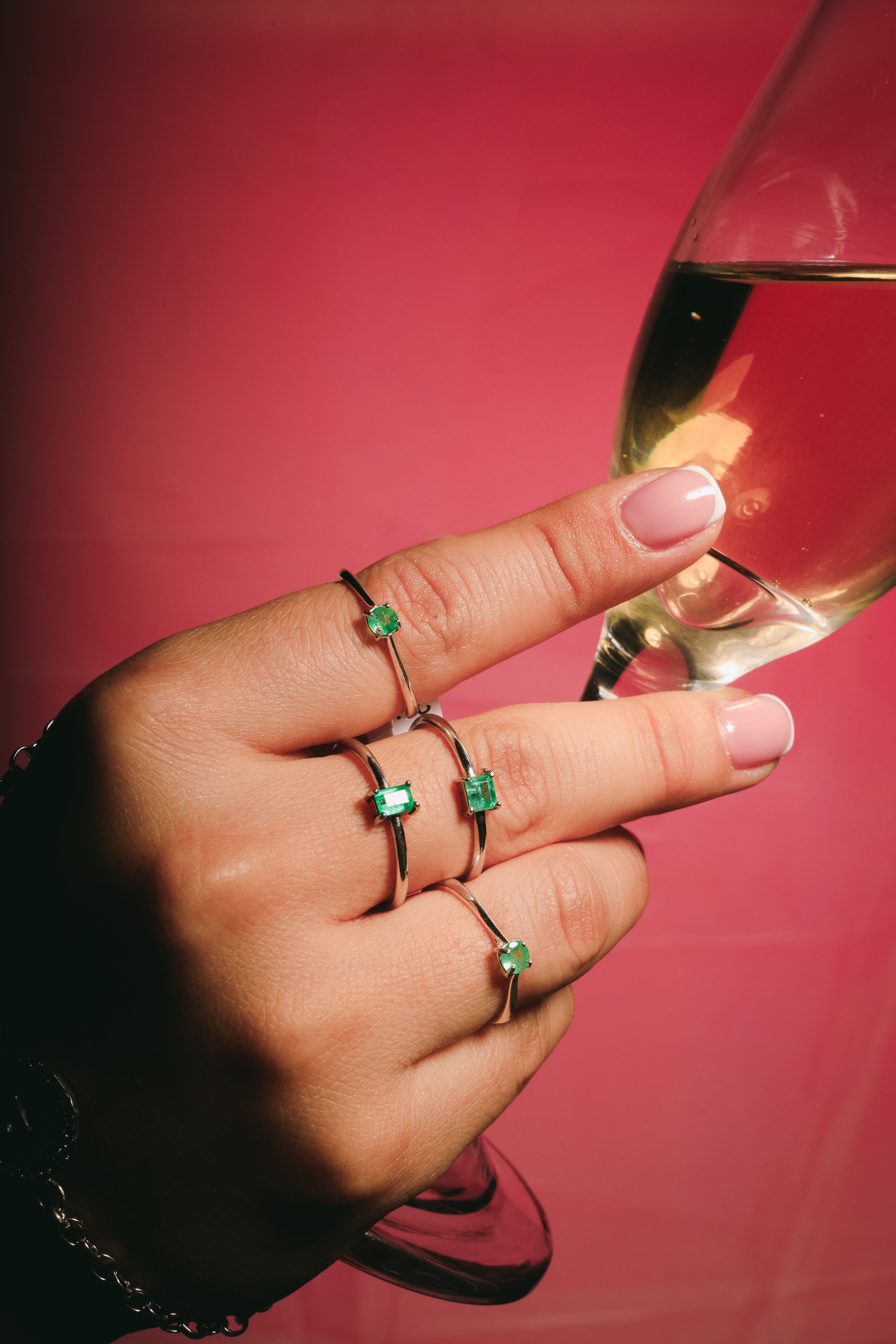 Stylish emerald rings on hand holding a champagne glass against pink background.