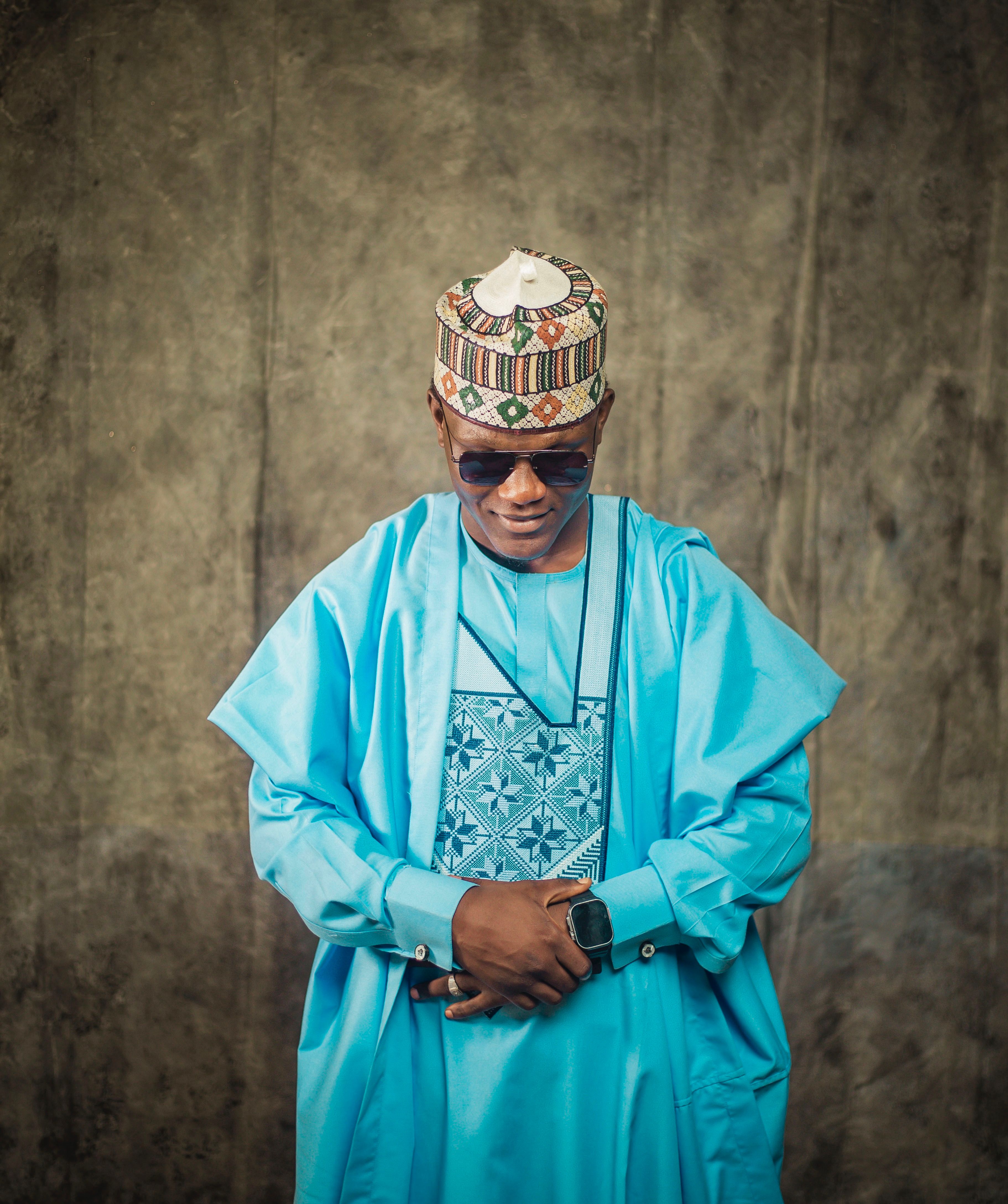Man in traditional blue agbada and cap with sunglasses poses against a textured backdrop.