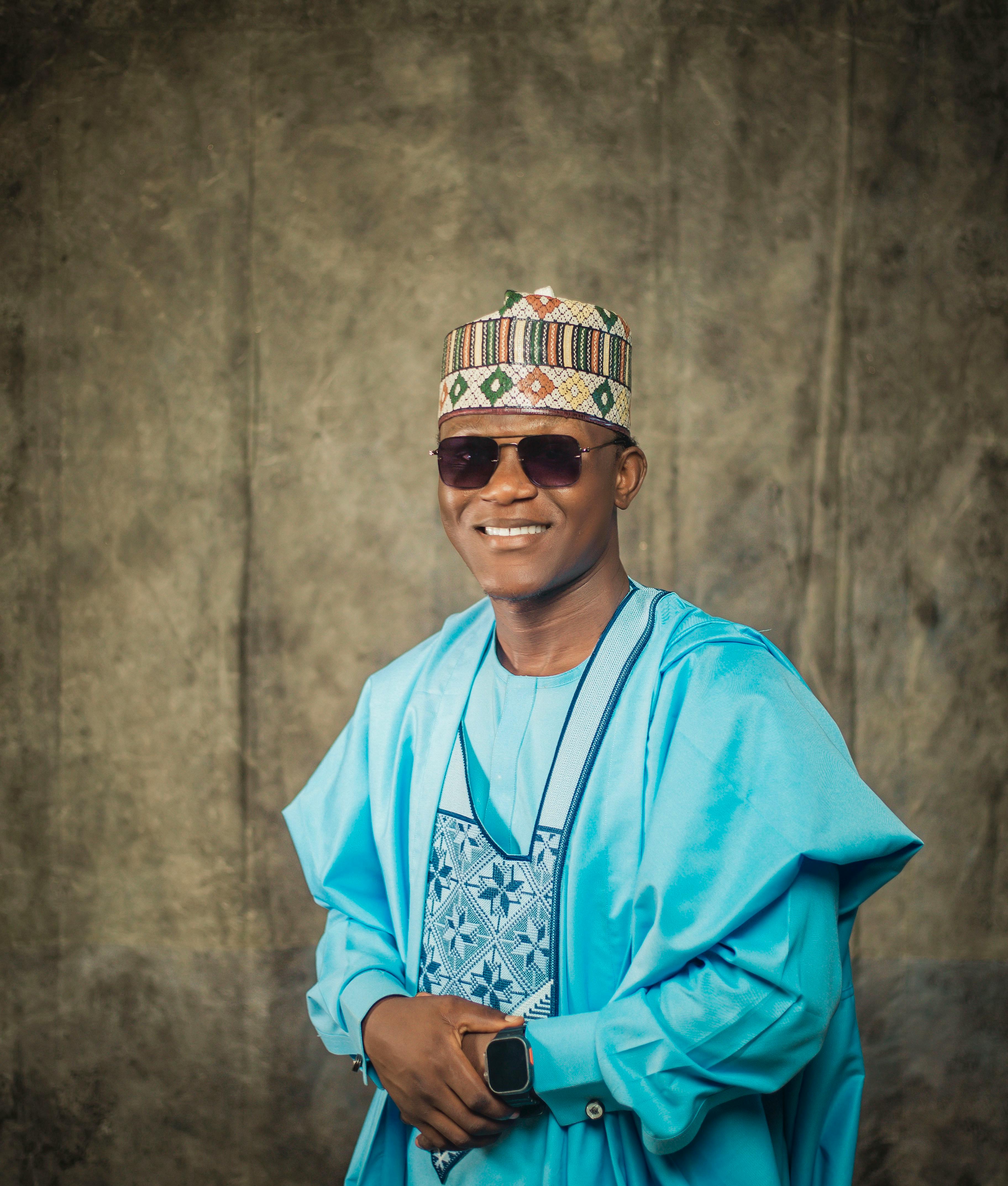Man in vibrant traditional Nigerian attire wearing sunglasses, smiling vividly.