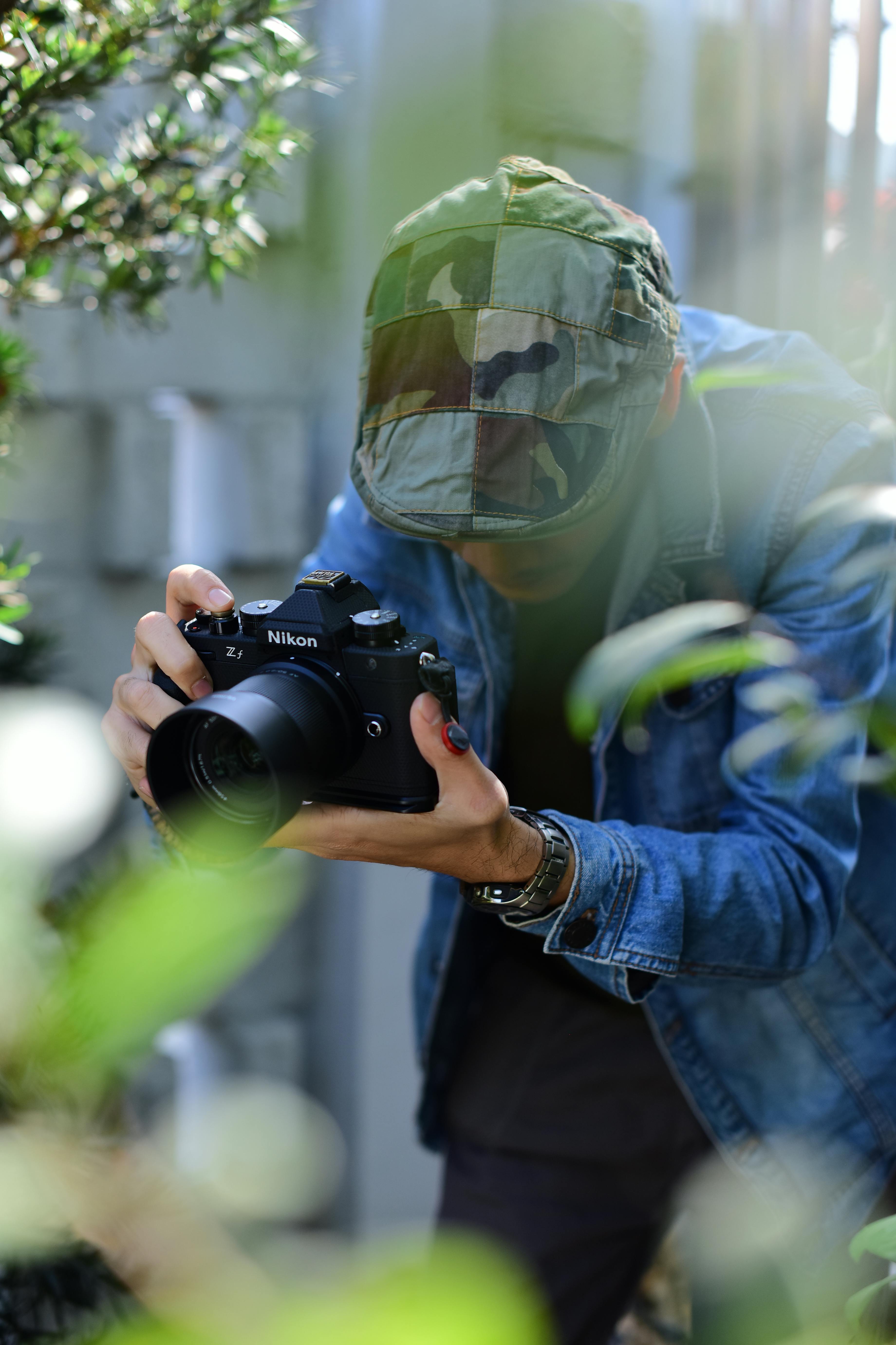 Free A photographer in a camo hat and denim jacket takes photos with a DSLR camera surrounded by greenery. Stock Photo