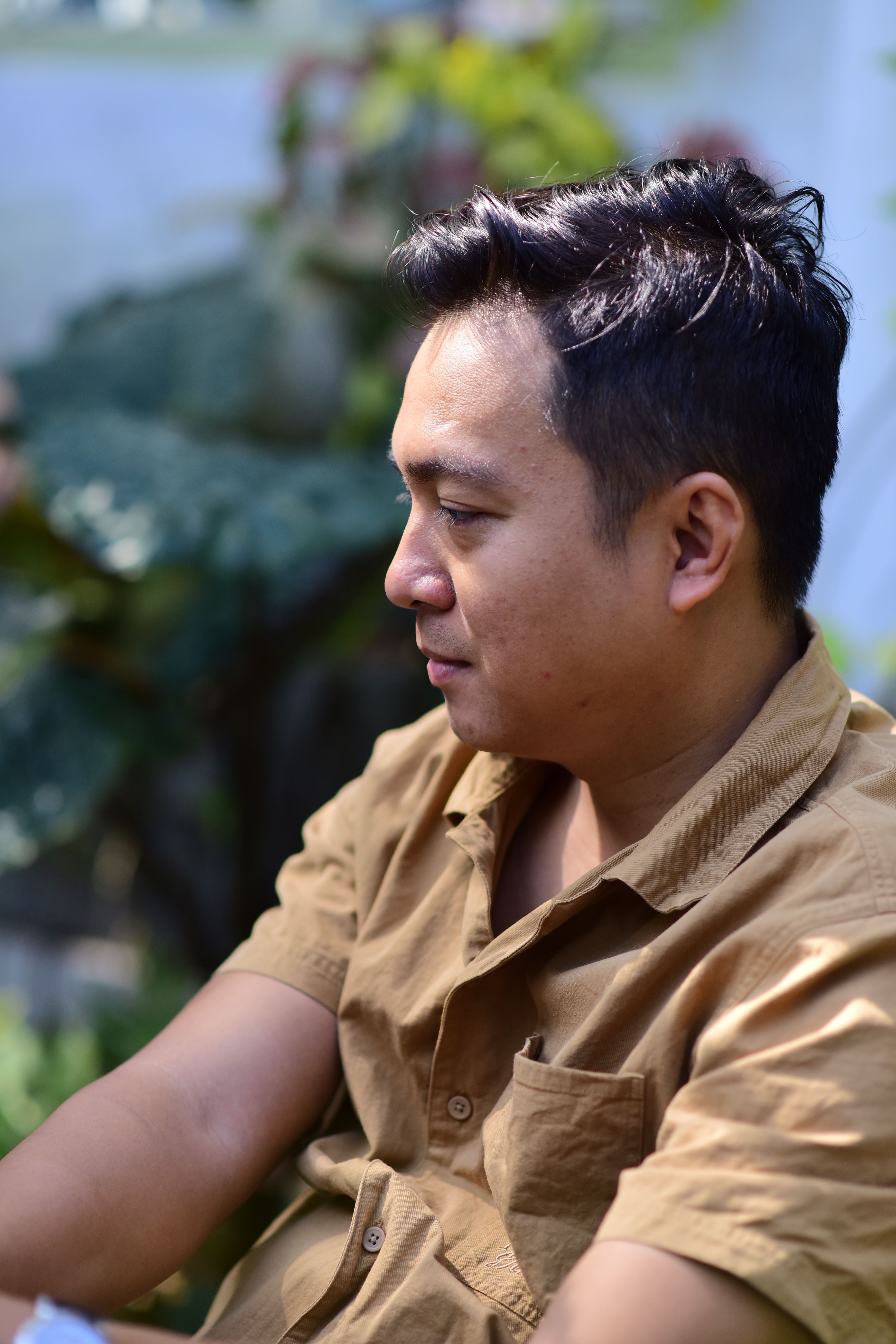 Casual portrait of a man in a brown shirt relaxing outdoors, surrounded by greenery.