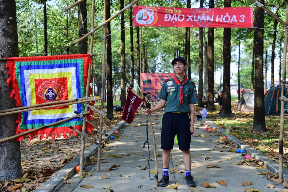 Boy scout standing under banner at outdoor camp with colorful flags and tents.