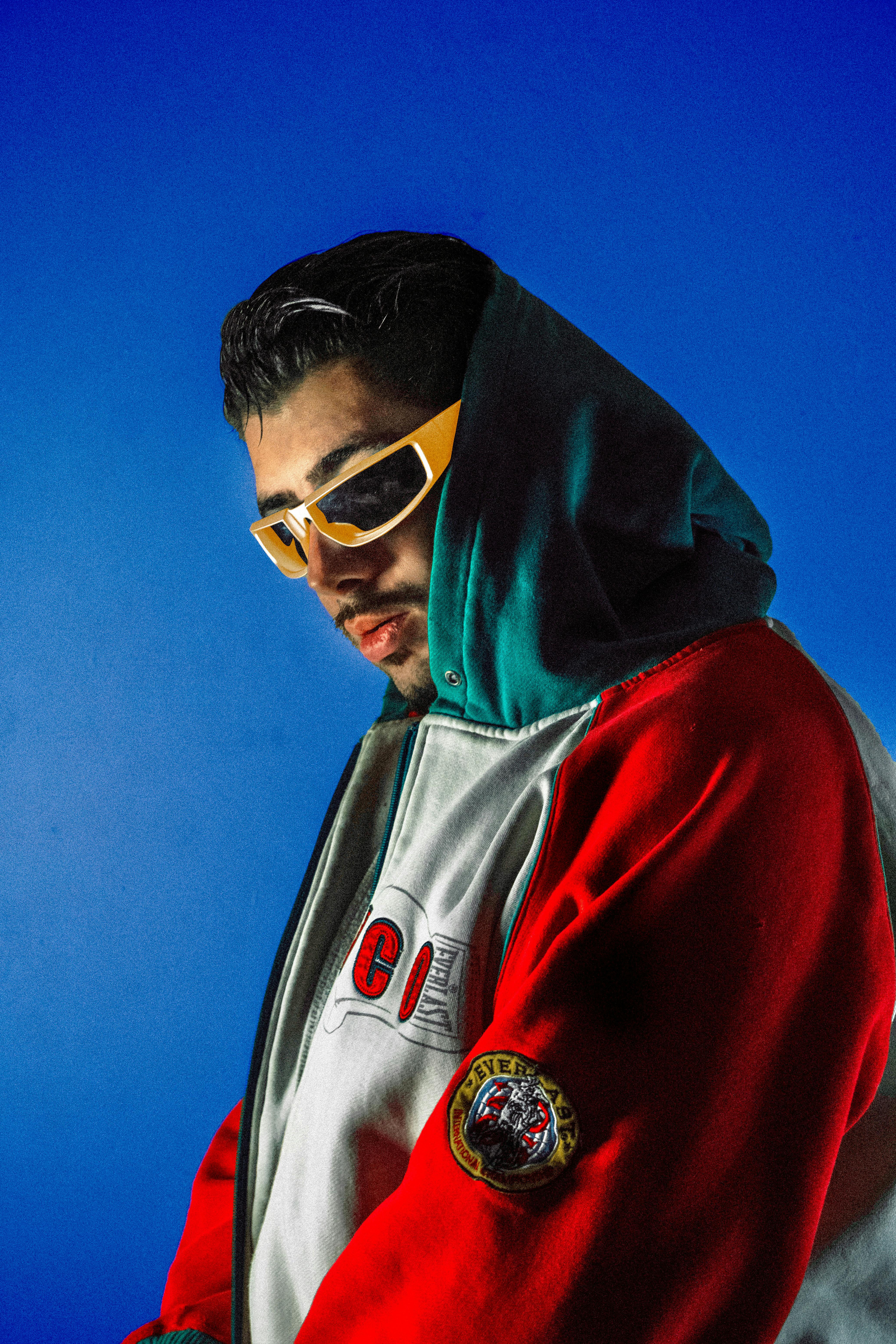 Fashionable man in hoodie and sunglasses posing in photo studio with blue background.