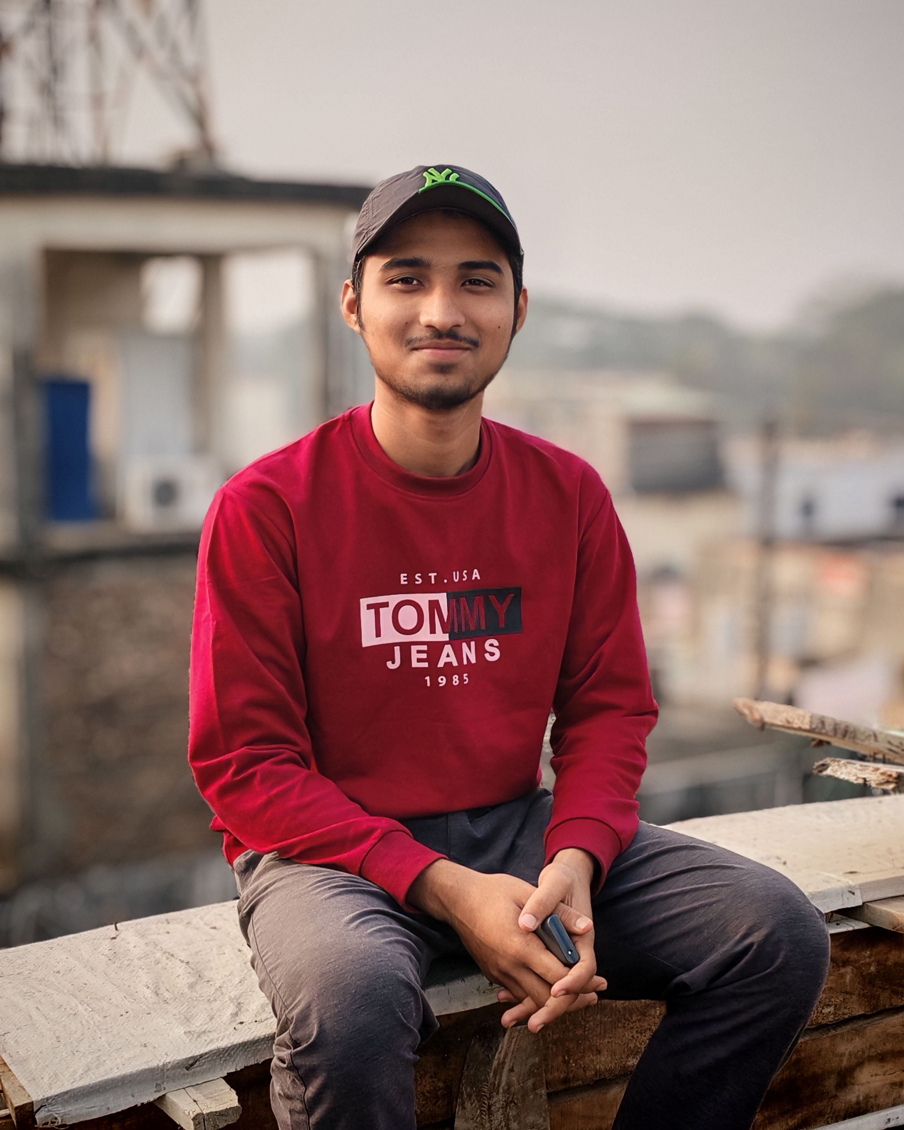 A smiling young man in a red sweatshirt, sitting outdoors on an urban rooftop.