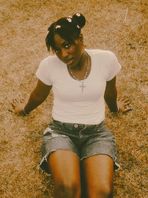 Free A young woman sitting on dry grass wearing a white shirt and denim shorts enjoying her time outdoors. Stock Photo