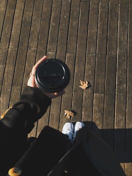 Overhead view of a person holding a coffee cup on a wooden deck with fallen leaves.