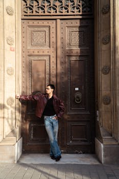 A stylish man in sunglasses and jacket leans against a large, detailed wooden door showcasing intricate design.