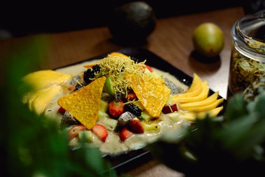 A vibrant fruit salad featuring mangoes, strawberries, papaya, nachos, and more, presented beautifully on a black plate.