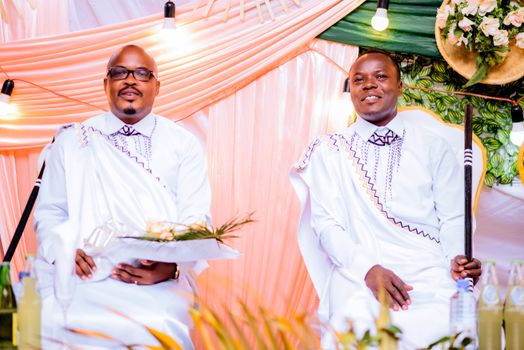 Two men in traditional white African attire seated at a ceremonial event indoors.