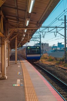 A blue commuter train arrives at an empty outdoor station on a sunny day.