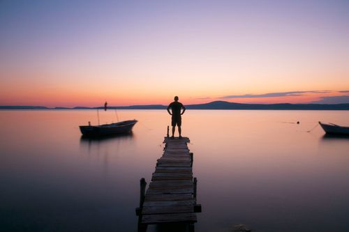 Free Lonely figure on a wooden pier overlooking calm water during a colorful sunset. Stock Photo