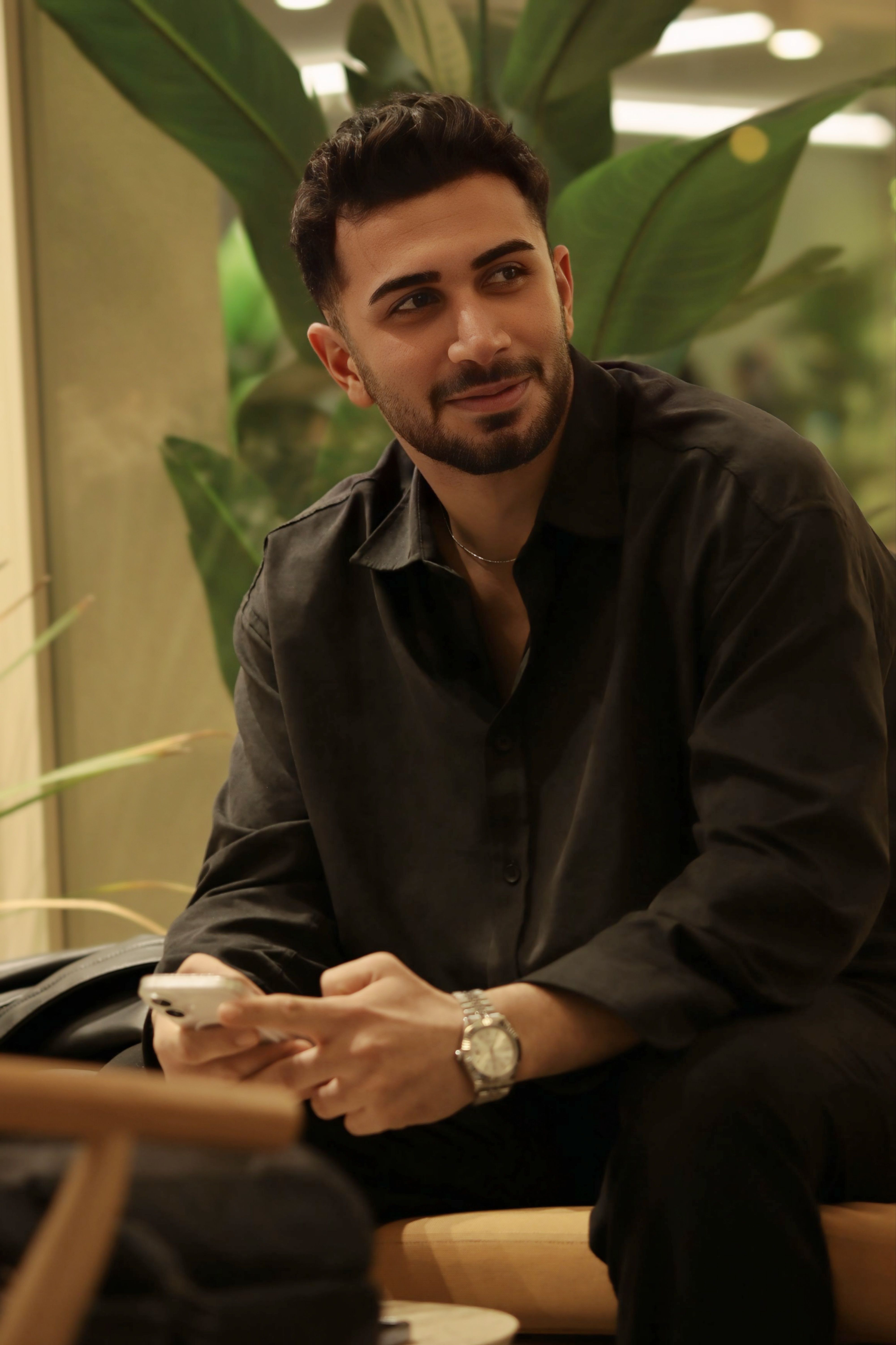Casual man in dark shirt sitting indoors holding smartphone, smiling warmly.