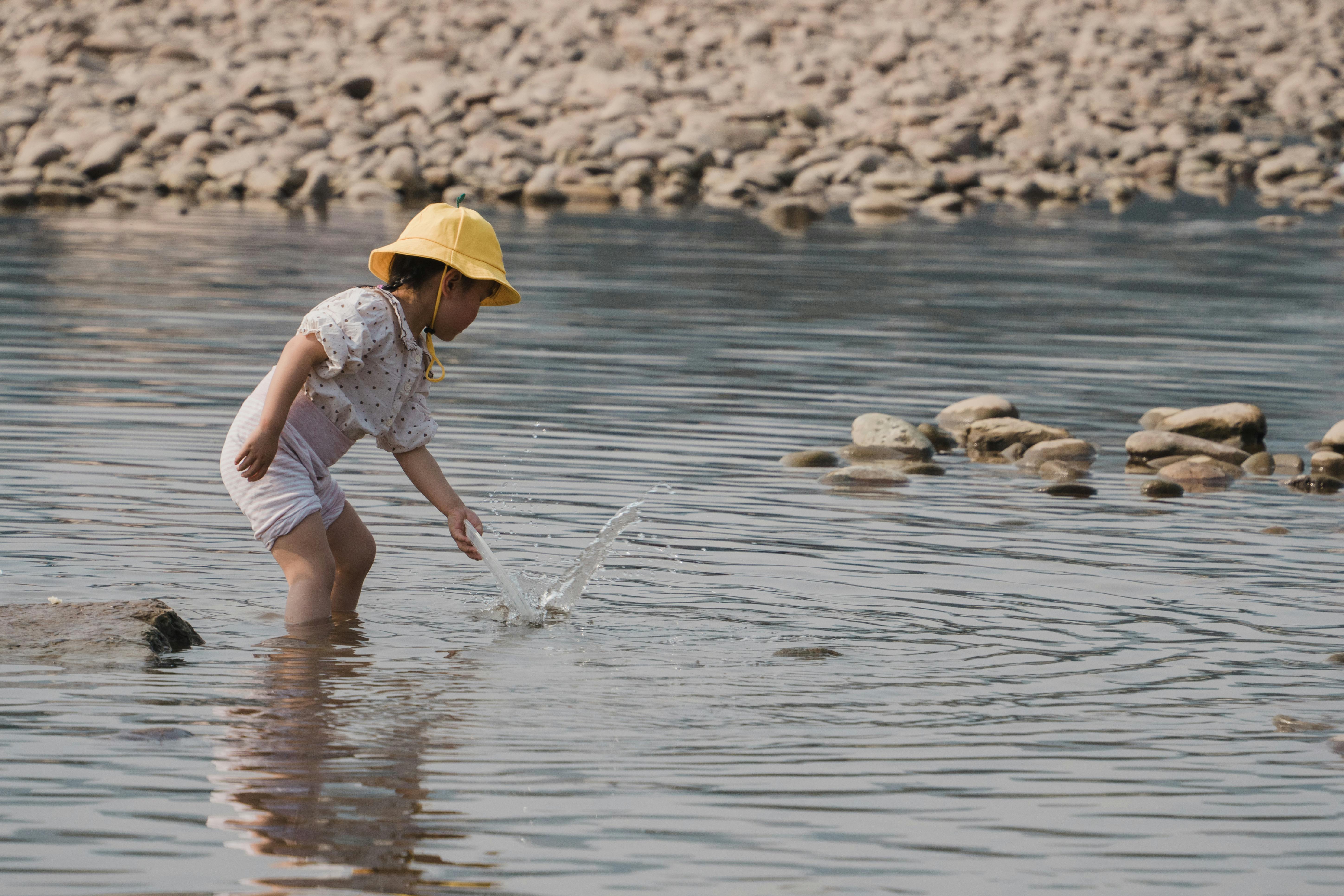A young child in a yellow hat plays joyfully at the edge of a river, splashing water.