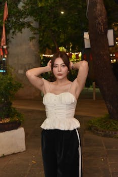 A young woman in a white top poses confidently outdoors at night with ambient lights creating a warm atmosphere.