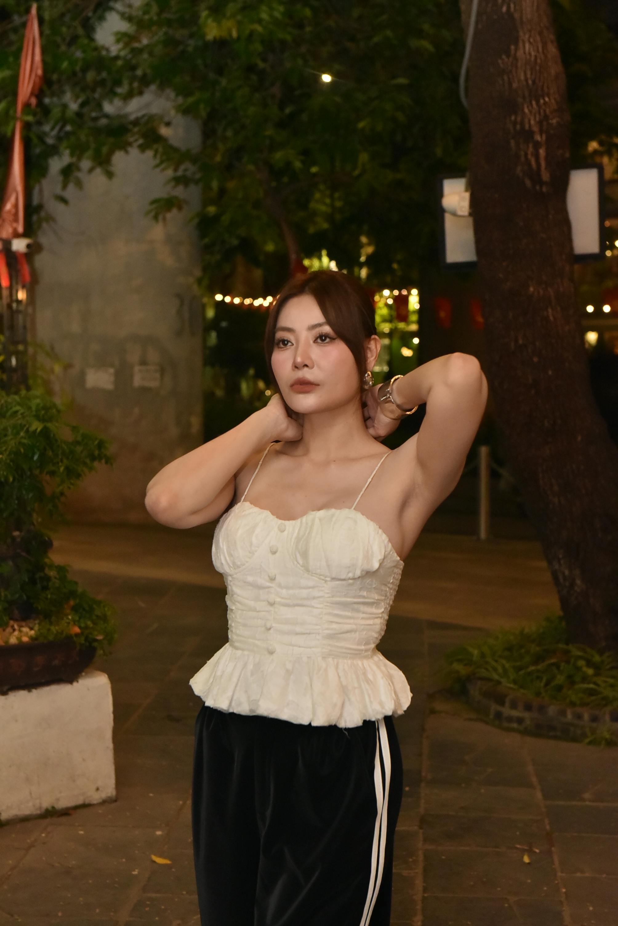 Gratis Una mujer elegante con un top blanco posa al aire libre por la noche en medio de luces urbanas y vegetación. Foto de stock