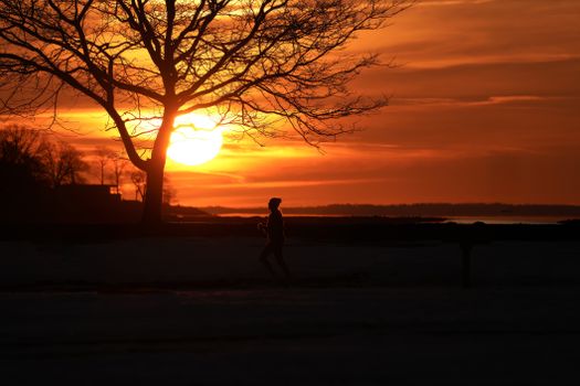 A jogger's silhouette against an orange sunset sky in a serene outdoor setting.
