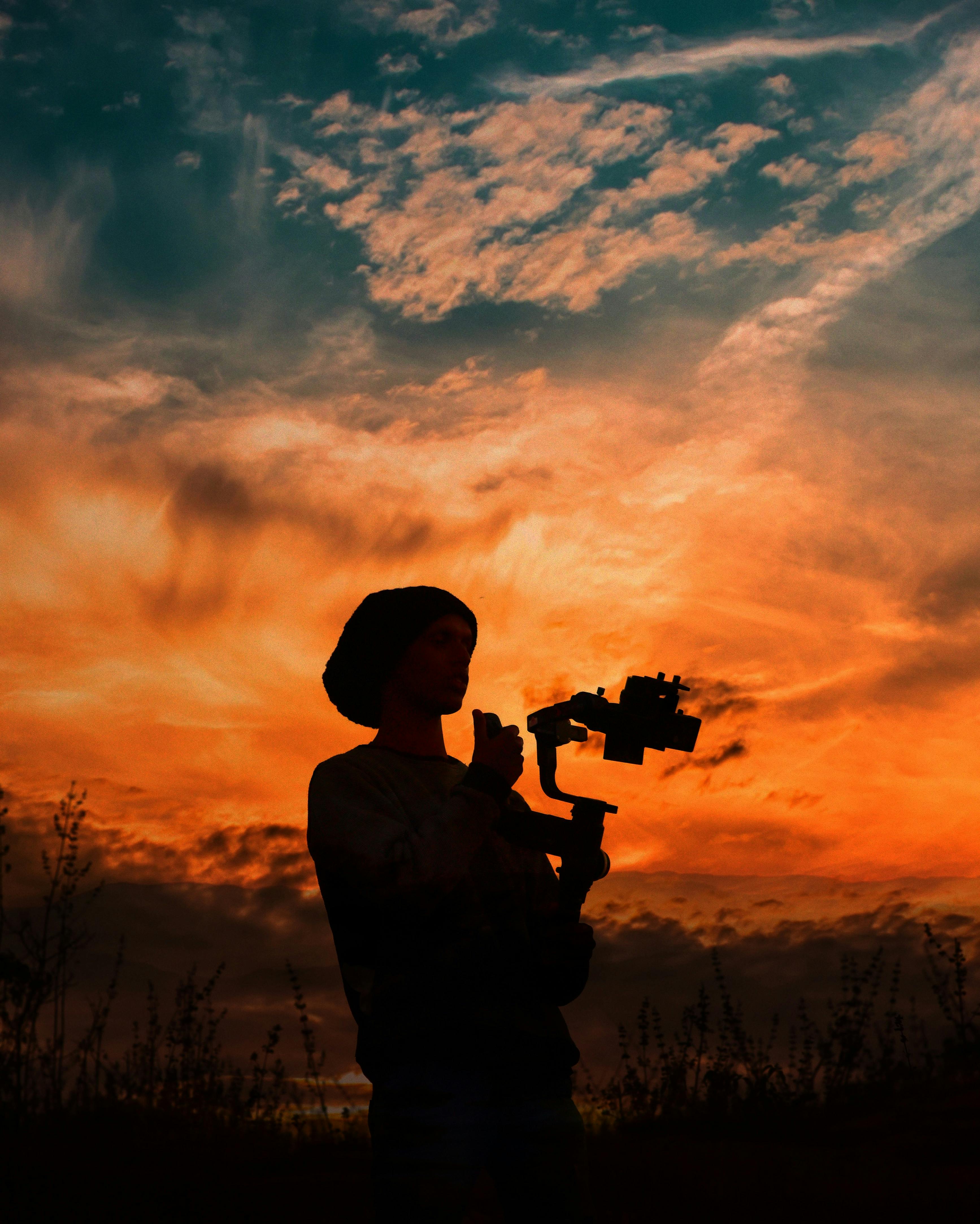 silhouette of person holding camera while standing under orange and blue sky