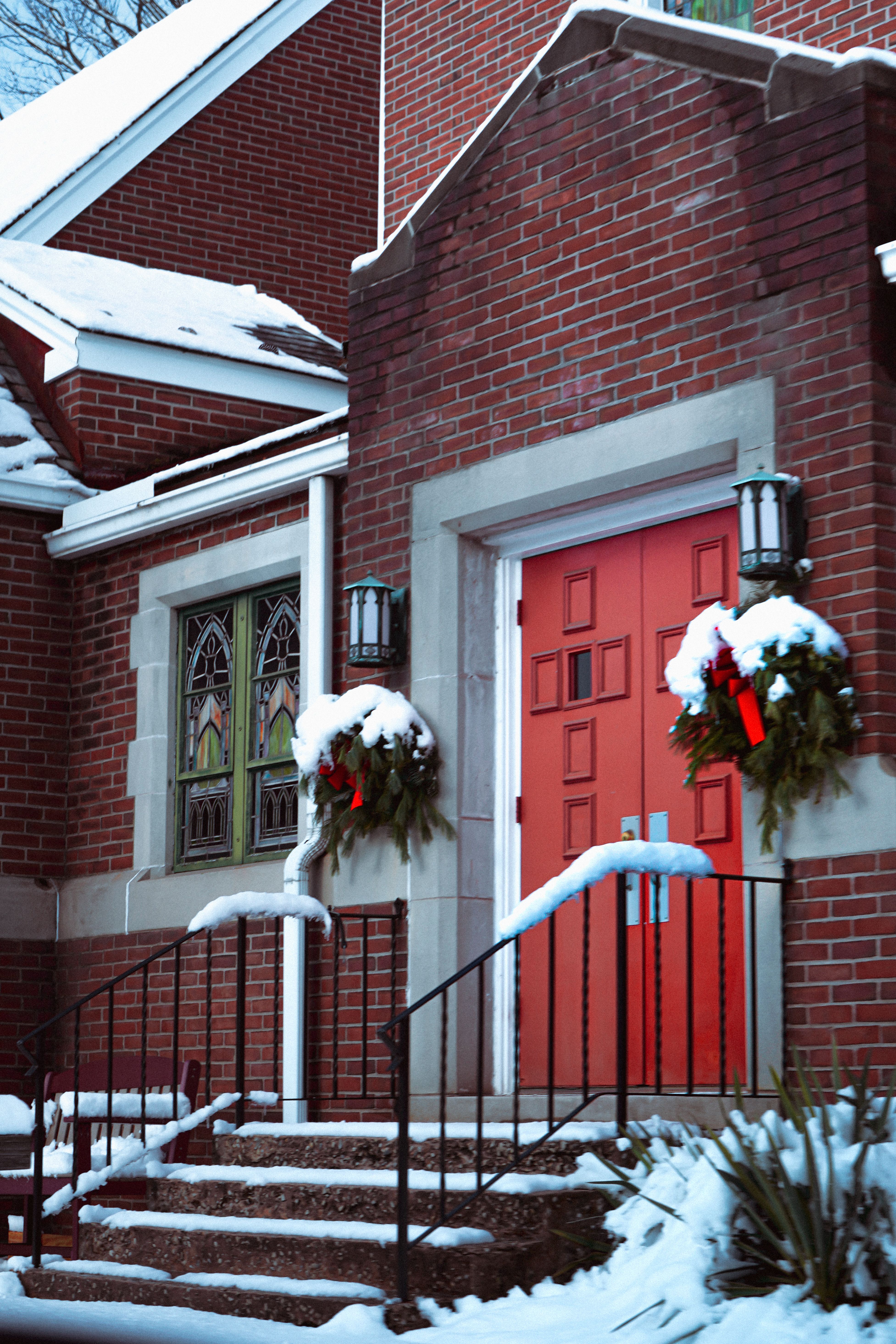 Snow-covered entrance of a brick church with red doors adorned by wreaths, symbolizing winter festivity.