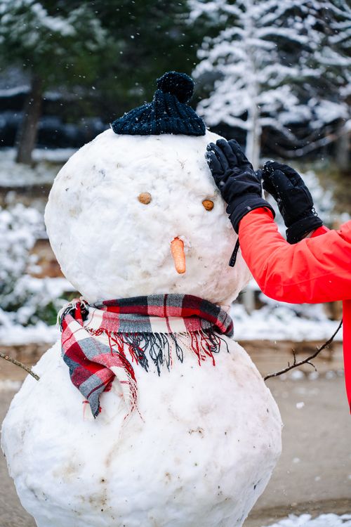 Free A person finishes building a cheerful snowman wearing a scarf and hat in a snowy outdoor setting. Stock Photo
