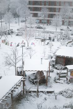 A snowy playground with children playing in the winter, surrounded by apartment buildings.