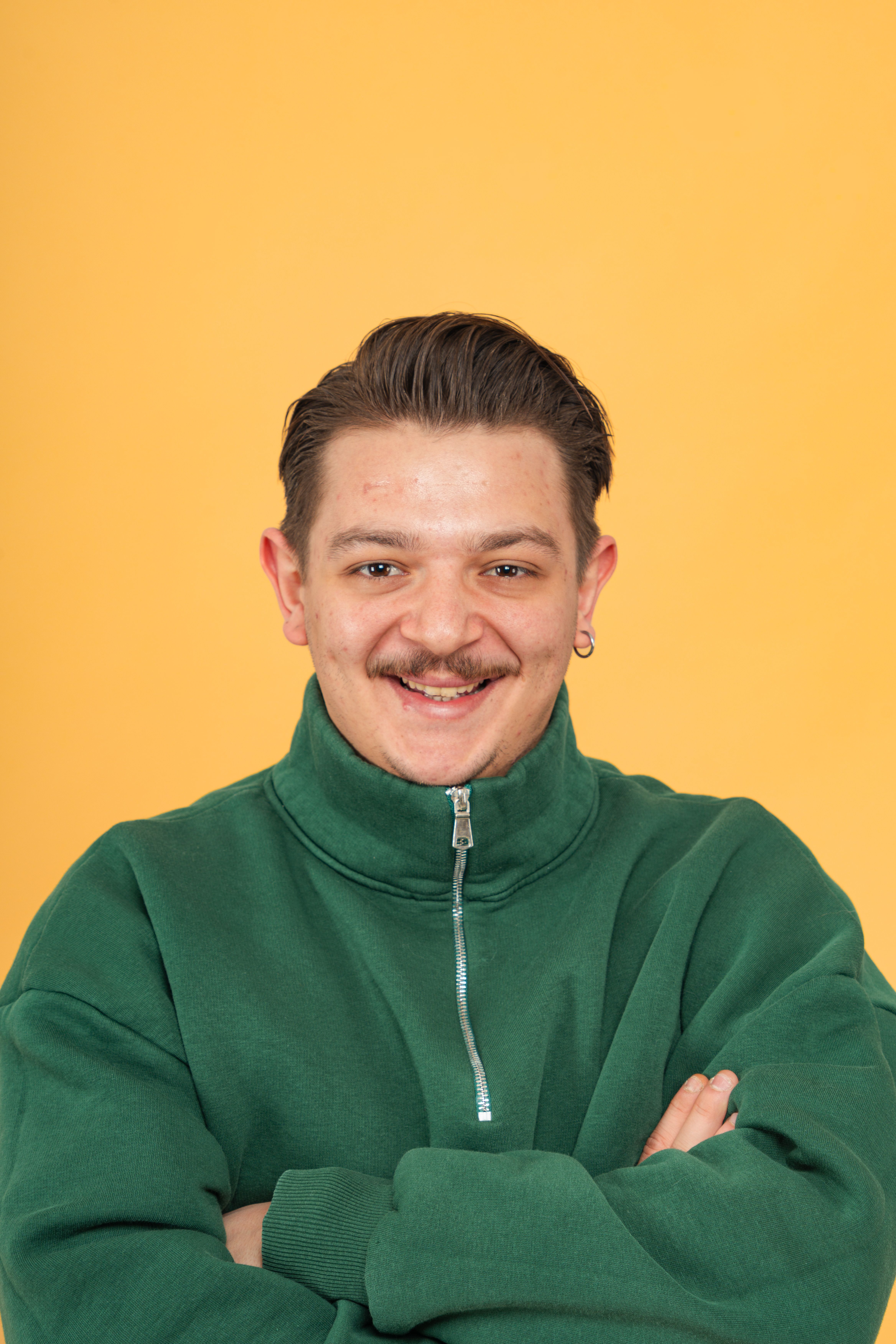 Portret van een lachende man in een groene jas met gekruiste armen tegen een felgele achtergrond.