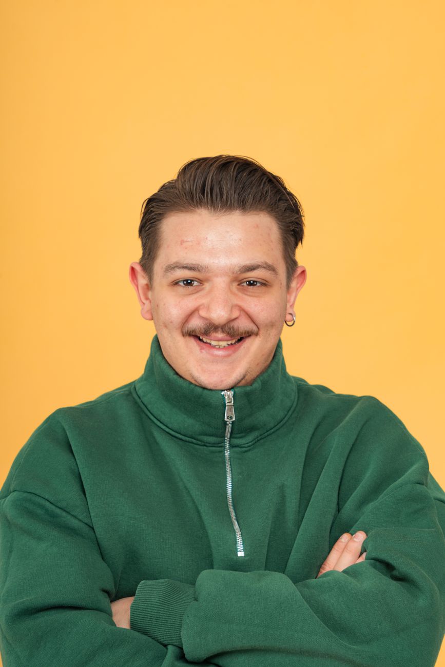Portret van een lachende man in een groene jas met gekruiste armen tegen een felgele achtergrond.